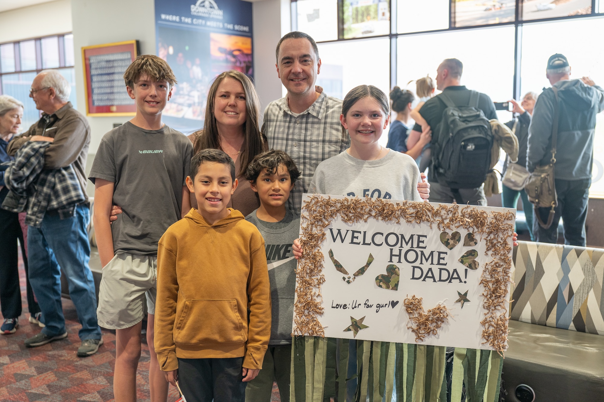 A family stands together for a photo, a girl holding a sign that reads "WELCOME HOME DADA!"