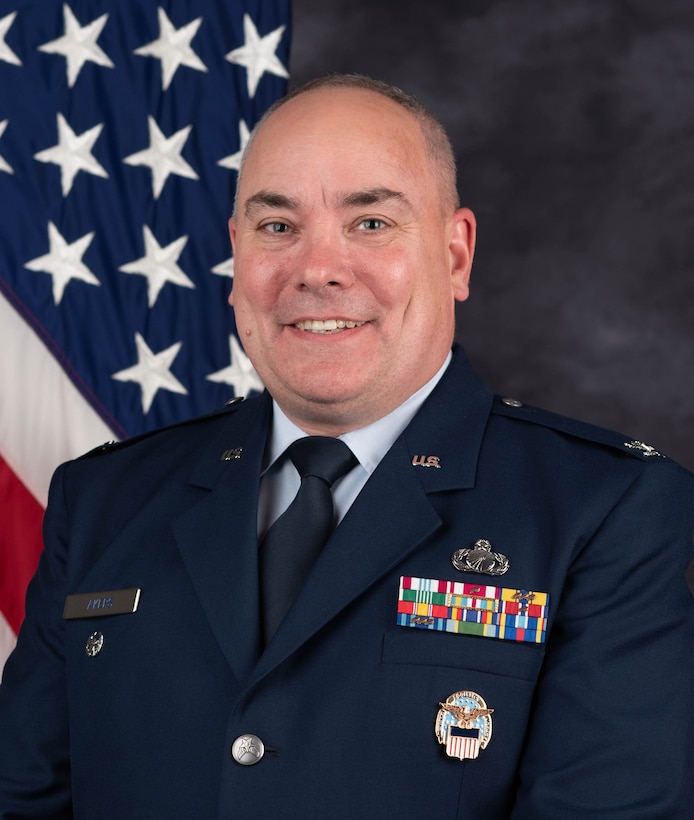 A smiling man in a dark blue Air Force dress uniform a flag positioned behind him.