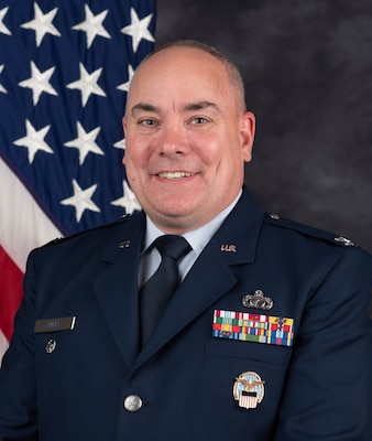 A smiling man in a dark blue Air Force dress uniform a flag positioned behind him.