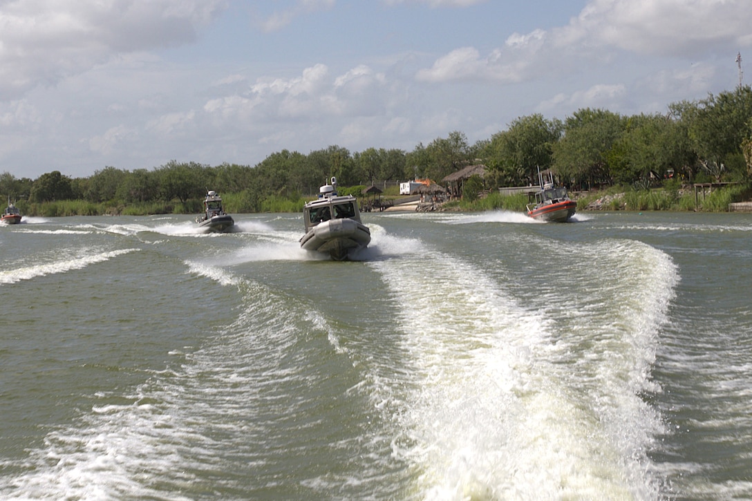 Four Border Patrol and Coast Guard boats cruise in a river on a cloudy day, with a tree-lined shore in the background.