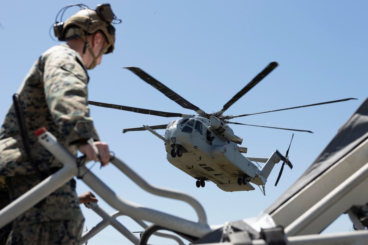 A Marine wearing a helmet holds a rail as a helicopter descends toward where the Marine is standing, with a blue sky in the background.