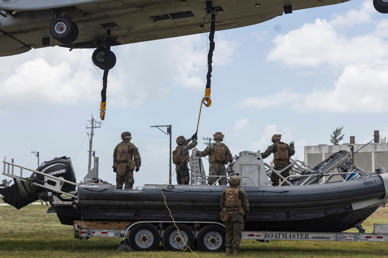 A Marine standing in a small boat uses a rod to guide a hook attached to a cable on a helicopter hovering above, as other Marines stand and watch, one standing outside of the boat, which is on a trailer.