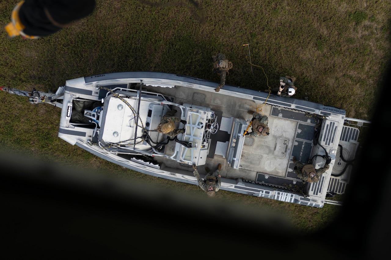 Four Marines stand in a small boat seen from above, with two others standing on the ground, as a hook on a cable is lowered from an open compartment of a hovering aircraft.
