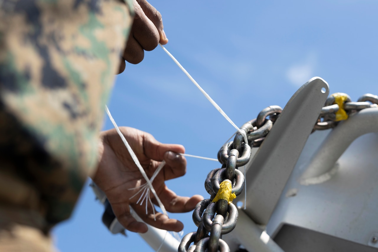 An unseen Marine ties a thin material to a chain attached to a larger object with another person standing nearby and a blue sky above.