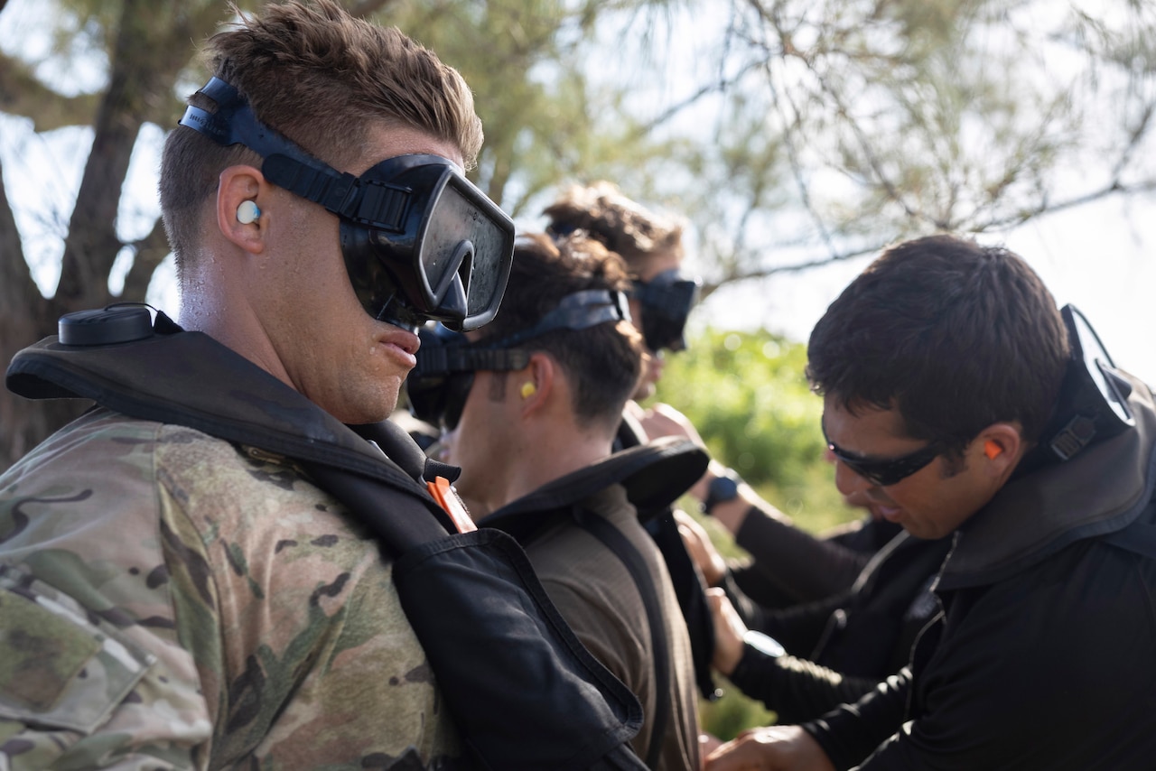 Soldiers wearing goggles and life vests line up outdoors as others help adjust equipment they're wearing, with tree branches in the background.