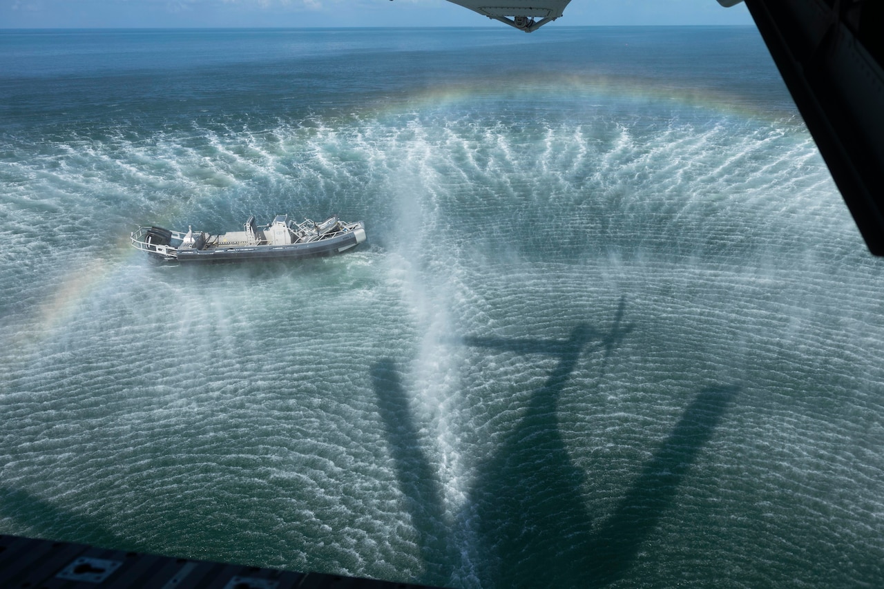 A boat is seen in rippling water from an open compartment of a hovering helicopter, its shadow reflected in the water, with a rainbow also visible.