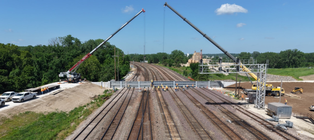 Two cranes lift grey structures over several brown railroad tracks with trees and blue sky in the background.