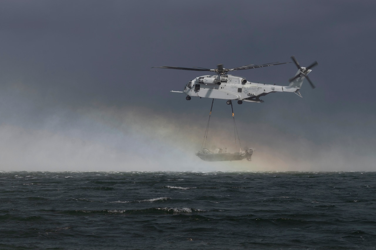 A helicopter with a boat attached to its underside by cables flies over a body of water, with a cloudy sky and rainbow in the background.