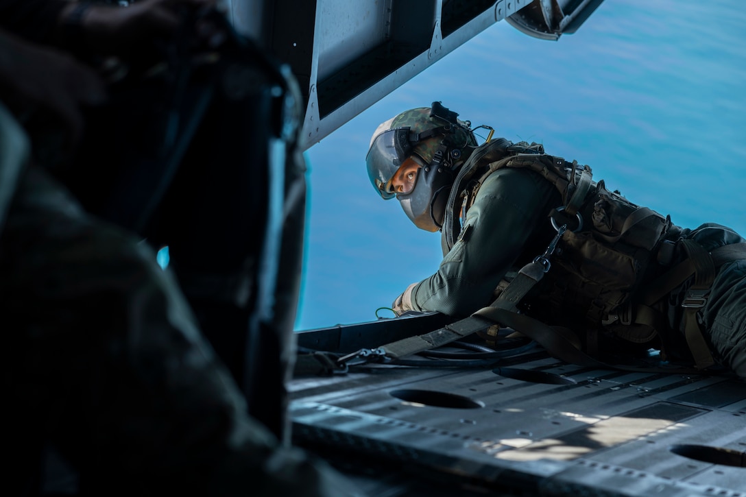 A Marine in a harness and helmet lays on the edge of an open aircraft in flight over water.