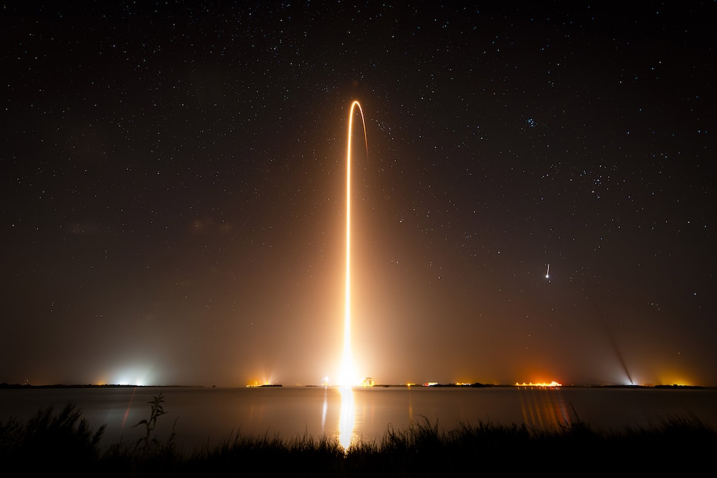 A rocket trail of light shoots up into the stars over the ocean at night.