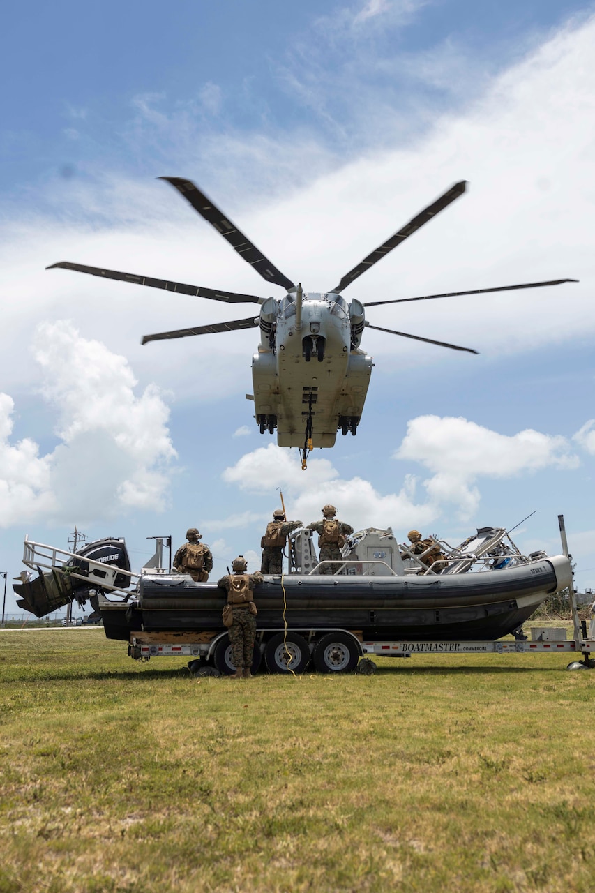 Four Marines stand in a small boat on a trailer on a grassy field, one holding a rod, as a helicopter descends toward the boat; another Marine stands on the ground, near the boat.