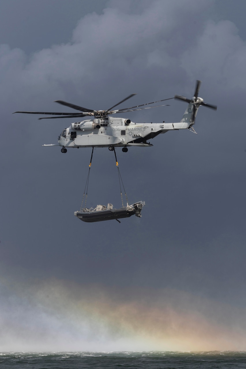 A helicopter with a boat attached to its underside by cables flies over a body of water, with a cloudy sky and rainbow in the background.