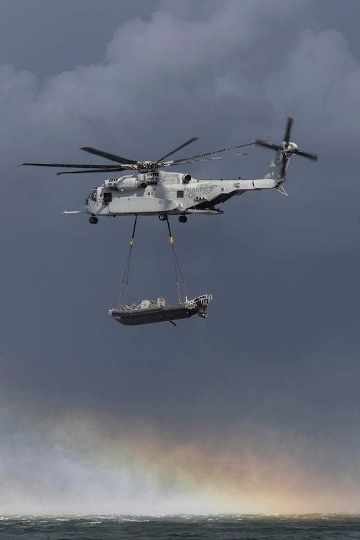 A helicopter with a boat attached to its underside by cables flies over a body of water, with a cloudy sky and rainbow in the background.