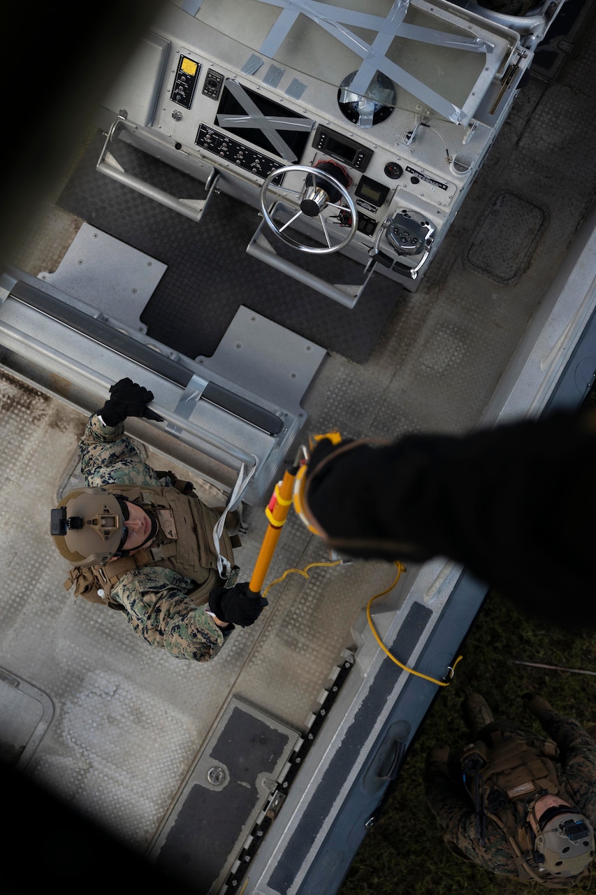 A Marine holds up a device to guide a cable descending from a hovering  aircraft toward a boat that the Marine is standing in.
