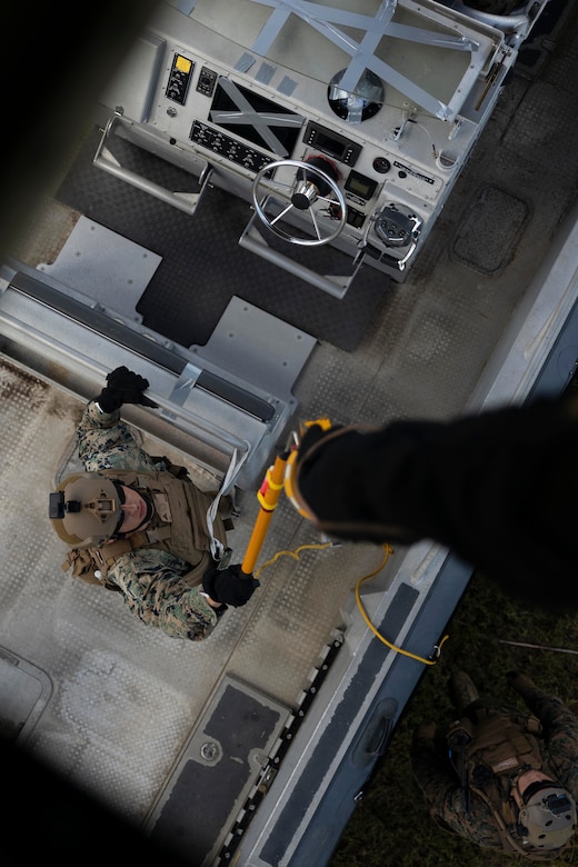 A Marine holds up a device to guide a cable descending from a hovering  aircraft toward a boat that the Marine is standing in.