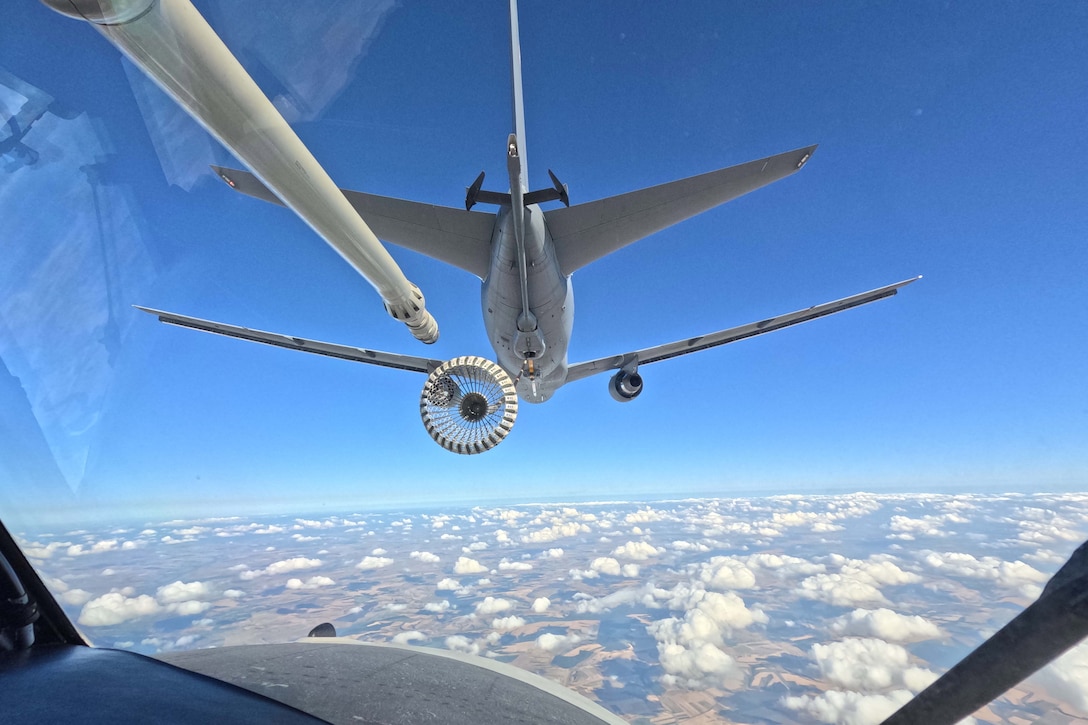 An aircraft flies above clouds in front of another aircraft, which positions a fueling tube towards the first aircraft.