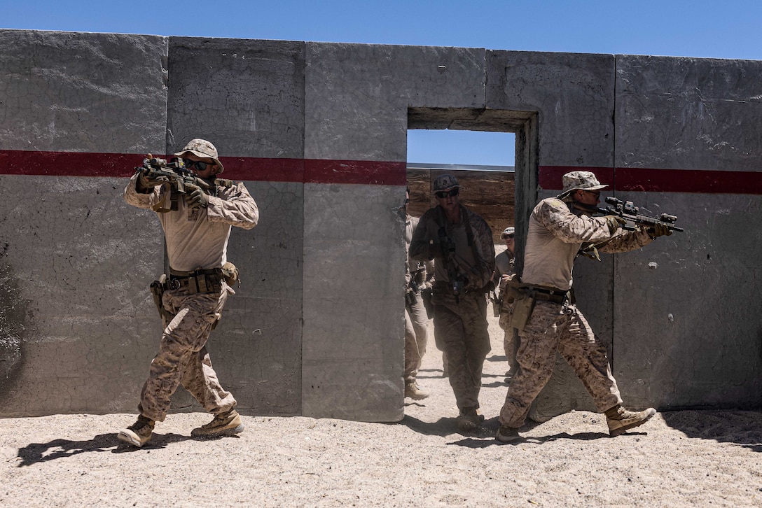 Two Marines with service weapons walk through a doorway as more Marines wait on the other side in an open-air training structure on a sunny day.