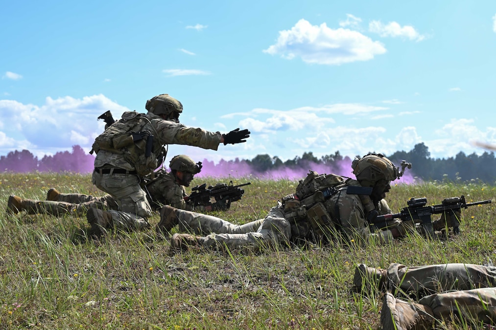 Soldiers lay in a field aiming service weapons while one crouches and gestures to the right with purple smoke and trees in the distance.