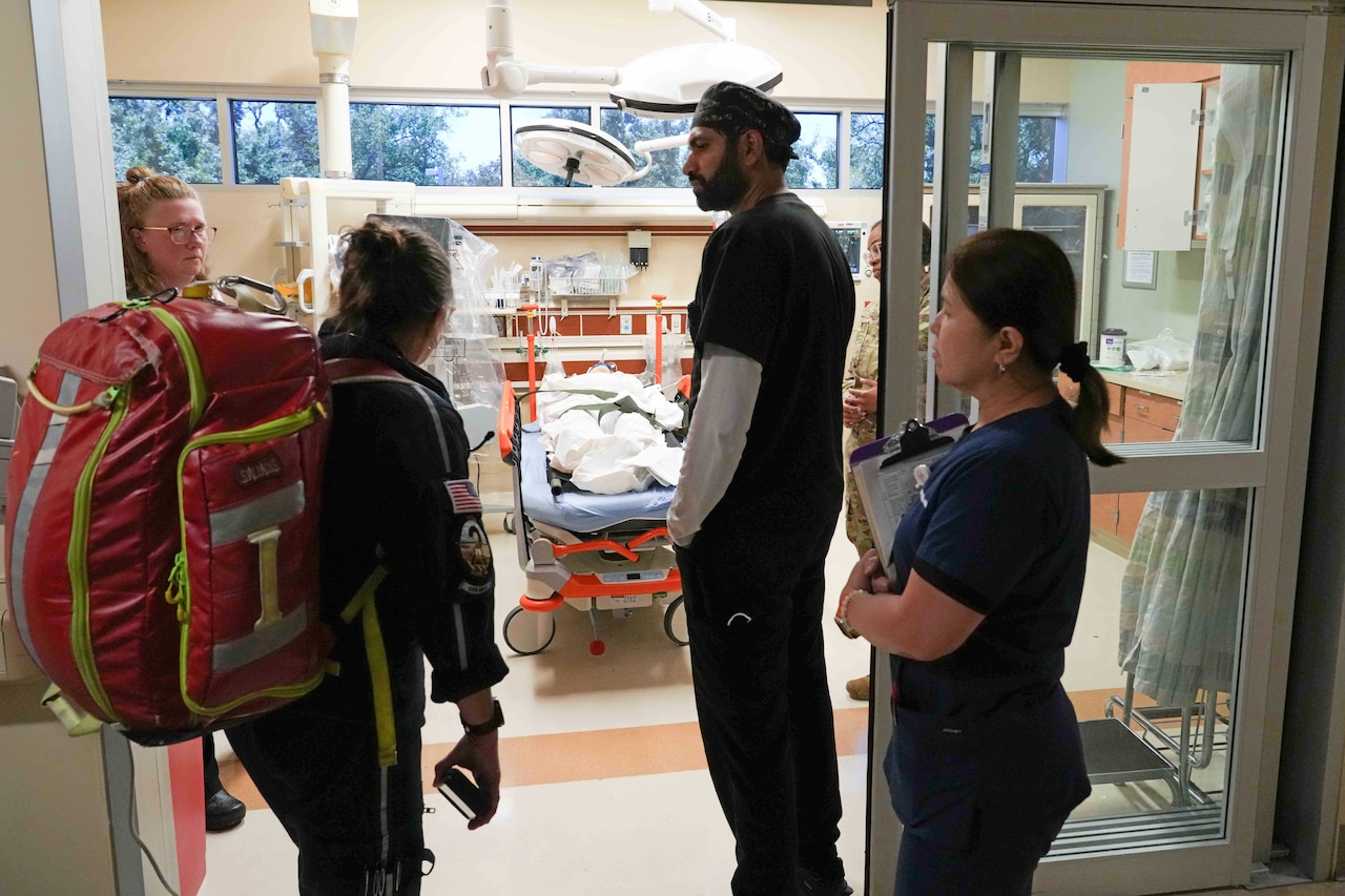 Medical personnel chat beside the door of a hospital room with a patient inside.