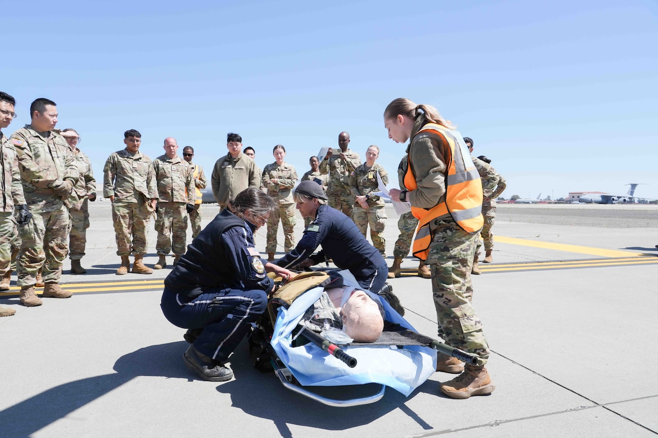 Medical personnel treat a patient lying on a gurney on a tarmac with troops in camouflage uniforms looking on.