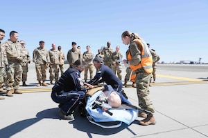Medical personnel treat a patient lying on a gurney on a tarmac with troops in camouflage uniforms looking on.