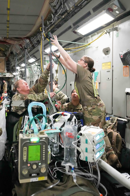 Troops in camouflage uniforms adjust medical equipment inside an airplane.