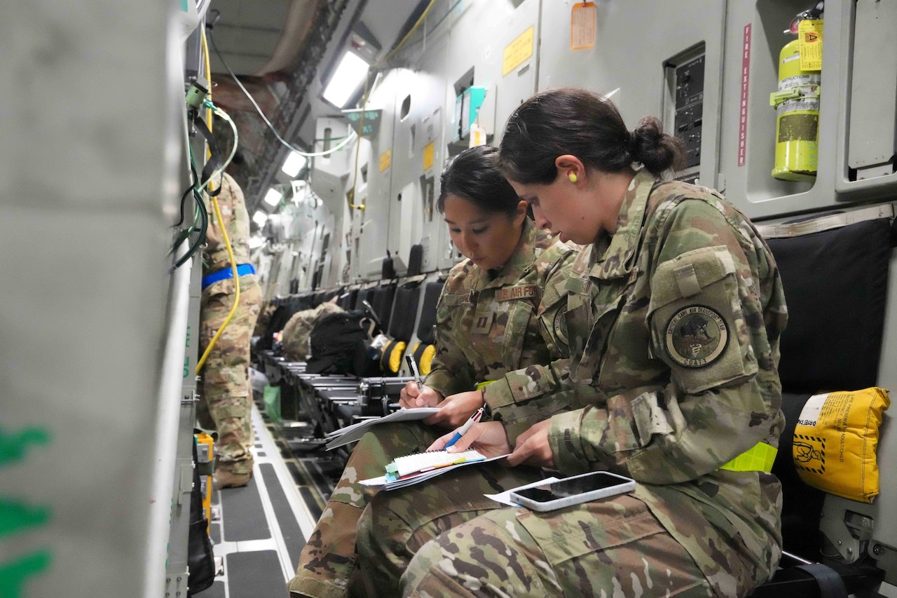 Two women dressed in camouflage uniforms, sit inside an airplane reviewing notes.