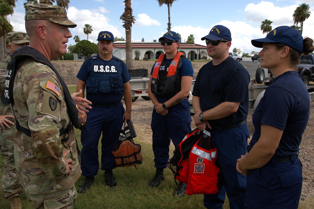 A soldier gestures while talking to four Coast Guardsmen as another soldier looks on outdoors, with a building, a wall and tress in the background.