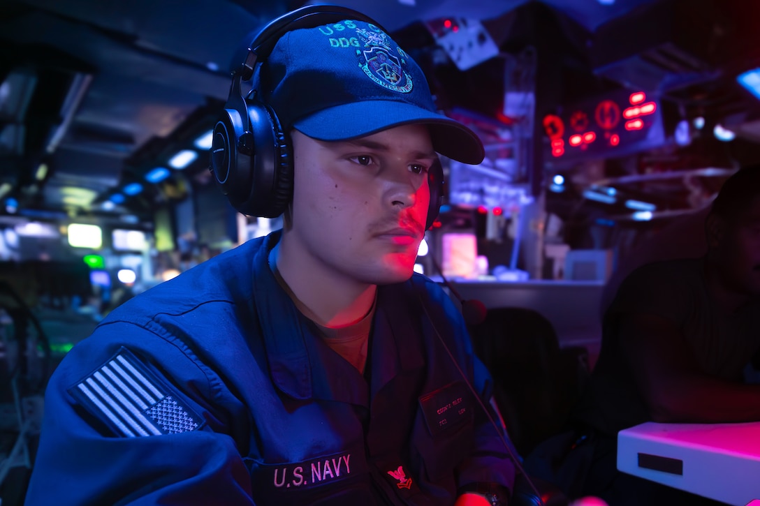 A sailor wearing a headset and cap monitors equipment in a control room setting of a ship.