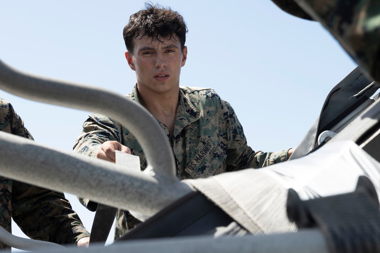 A Marine adjusts straps on a piece of equipment outdoors, with a blue sky in the background.