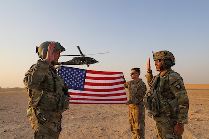 Two soldiers face each other and raise their right hands while two soldiers hold an American flag and a helicopter hovers in the background.