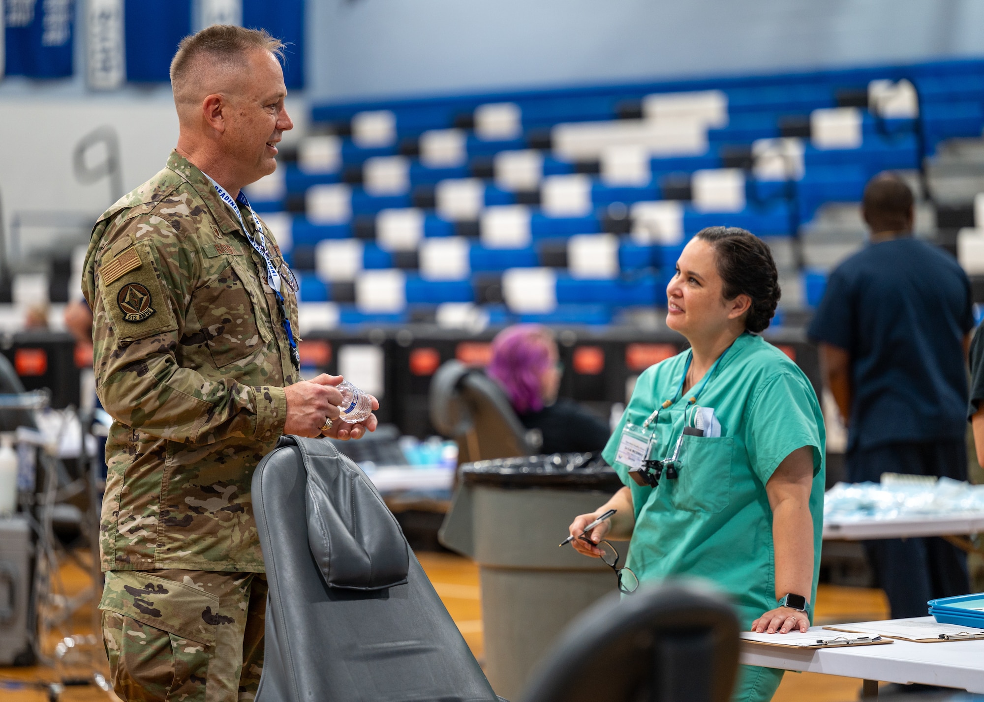A man in a military uniform talks to a woman in scrubs in a high school gym.