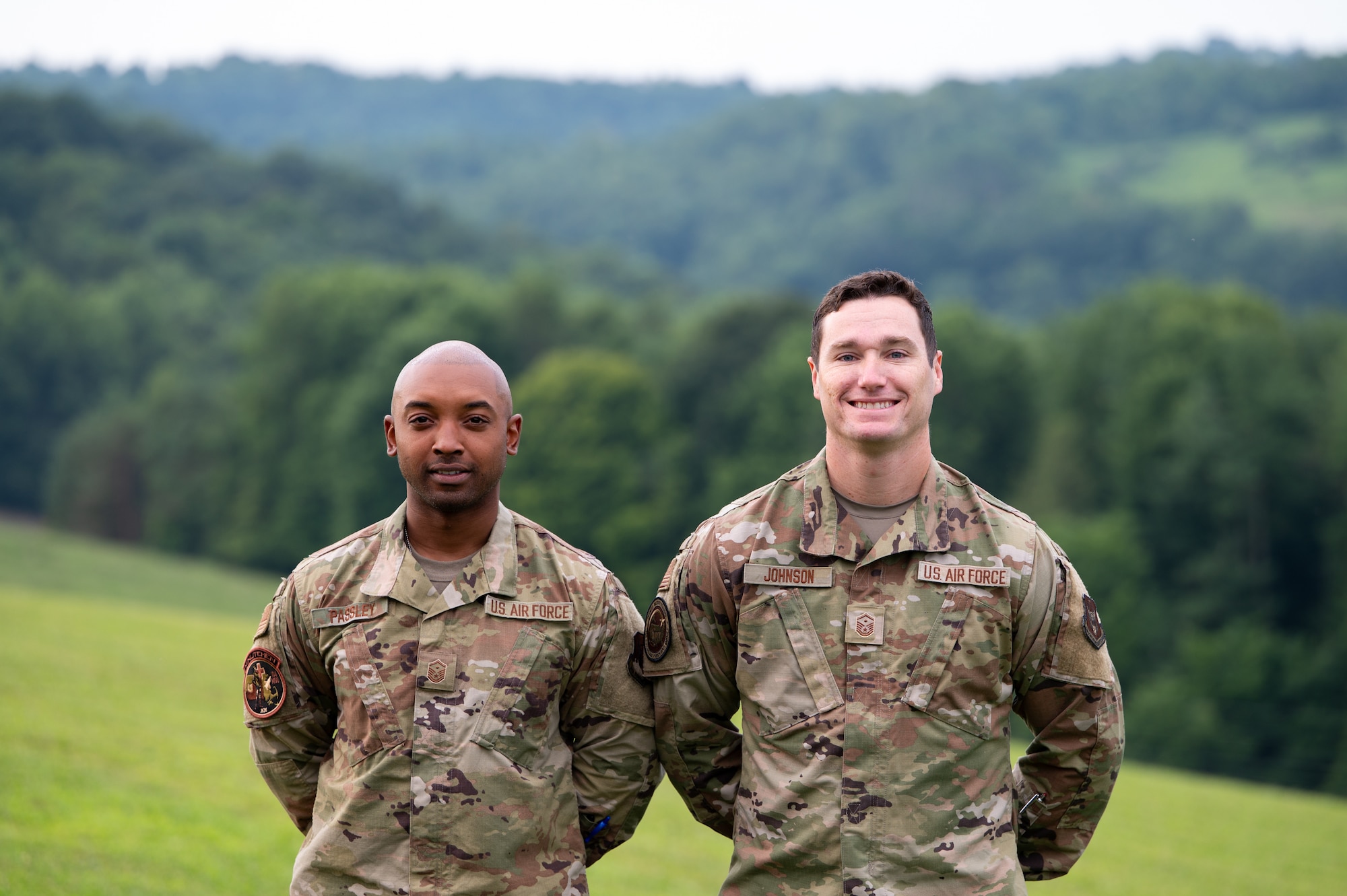 Two men in uniform smile while posing for a photo on a hillside.