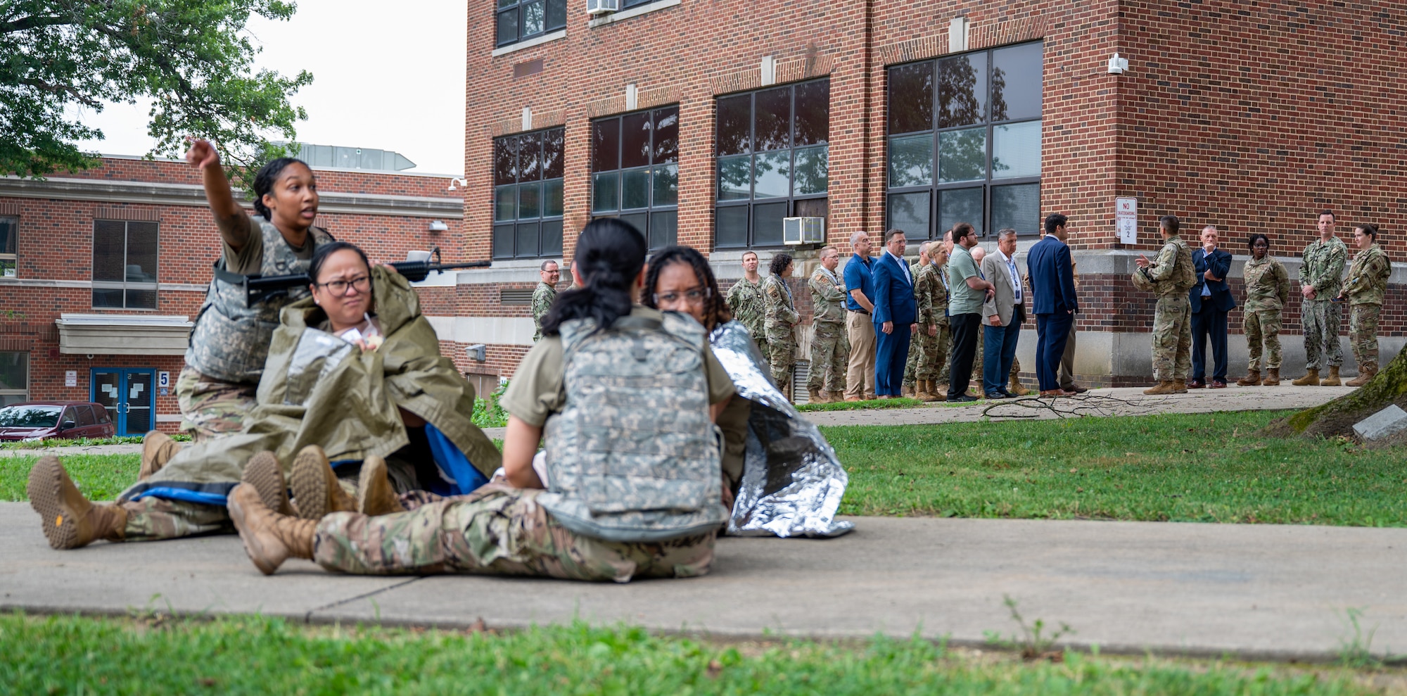 Service members complete military medical training as a group of spectators watch.