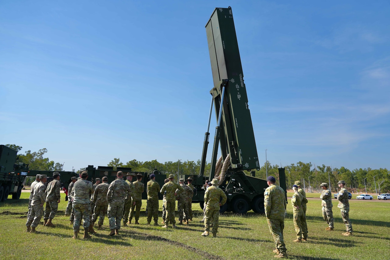 More than two dozen people in camouflage military uniforms stand in a field around a large military weapons system that is pointing in the air.