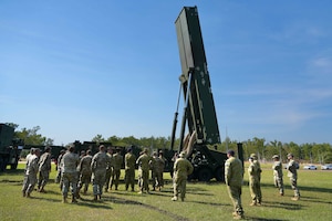 More than two dozen people in camouflage military uniforms stand in a field around a large military weapons system that is pointing in the air.