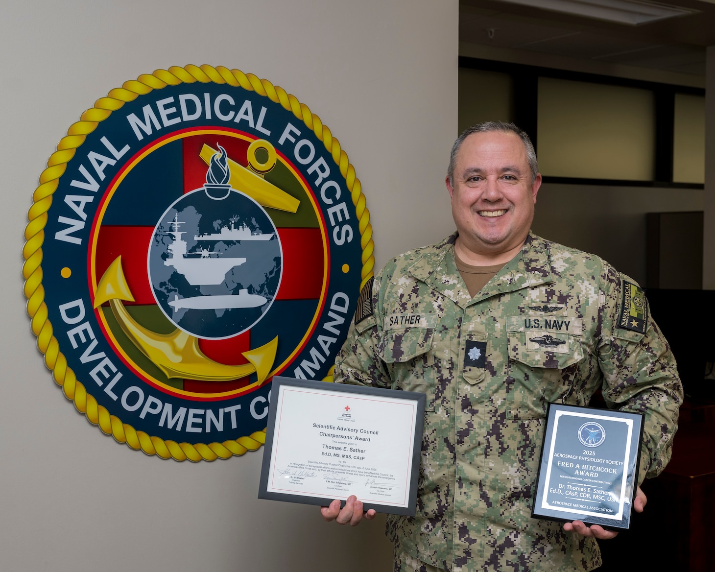 Cmdr. Thomas Sather, director of the Naval Medical Forces Development Command’s Tactical Combat Casualty Care program, displays his recent civilian awards at Joint Base San Antonio-Fort Sam Houston, Texas, Aug. 1, 2025. Sather was presented the American Red Cross Chairpersons’ Award for his significant contributions to the Red Cross Scientific Advisory Council, and recognized the Aerospace Medical Association with the Aerospace Physiology Society’s Fred A. Hitchcock Award for his outstanding performance as a senior aerospace physiologist. (U.S. Navy photo by Malcolm McClendon)