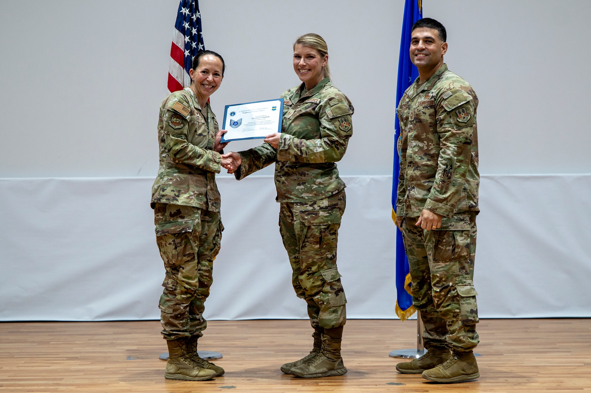 SSgt Campbell in the middle shaking Col. Quidley's hand to her left while both holding a certificate of promotion with Chief Correa to Campbell's right with flags in the background