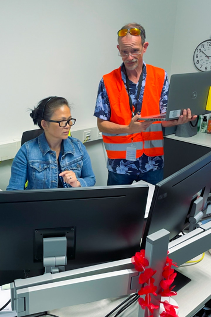 A man wearing a reflective safety vest and holding a laptop computer looks over the shoulder of a seated woman at a computer screen, which is sitting on her desk in front of her.