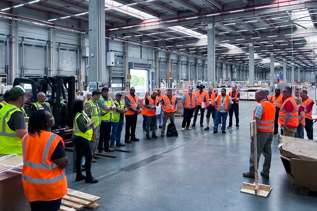 A large group of people wearing reflective safety vests stand around a warehouse looking at a person who is speaking to the group.