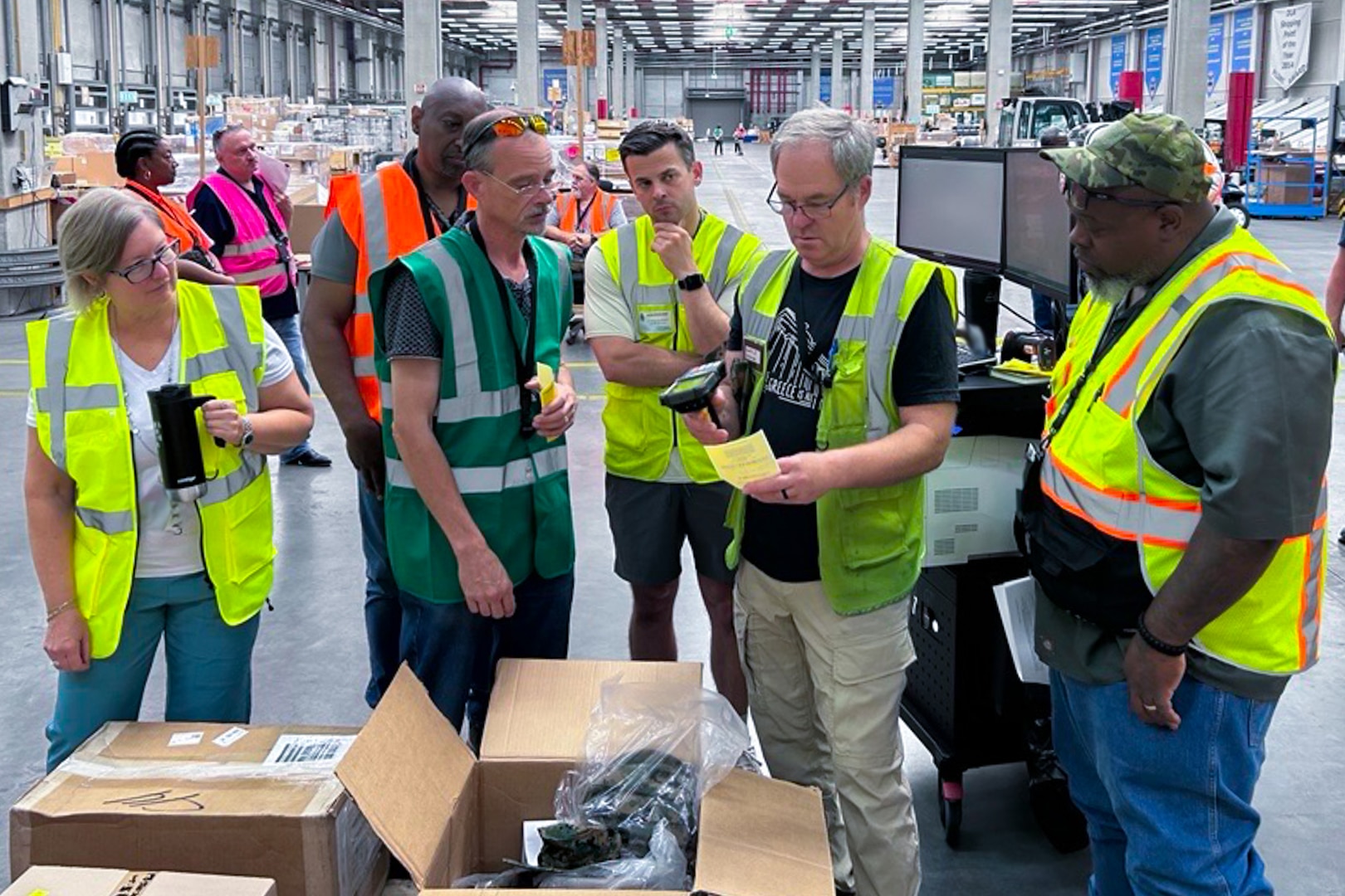 A group of people wearing reflective safety vests gather around a man scanning a ticket in a warehouse.