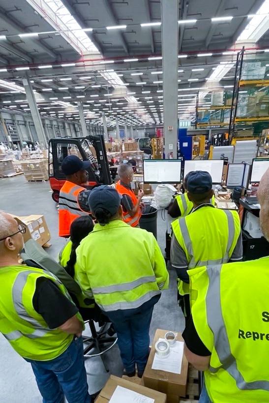 People wearing reflective safety vests gather in a group to watch a demonstration in a warehouse.