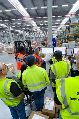 People wearing reflective safety vests gather in a group to watch a demonstration in a warehouse.