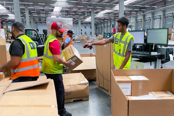 Men wearing reflective safety vests move brown boxes in a warehouse.