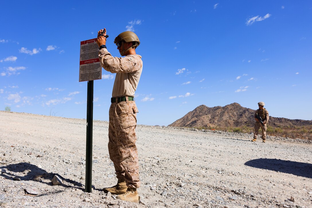 A Marine uses a tool to secure a sign to a post in a desert-like area as another Marine looks on, with a hill and blue sky in the background.