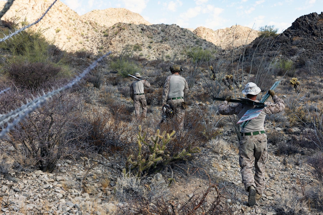 Marines carry signposts and other equipment in a hilly, rocky and sparsely vegetated area on a sunny day.