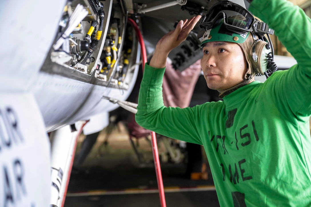 A sailor wearing protective gear kneels next to an oxygen generation system on an aircraft in a ship's hangar bay.