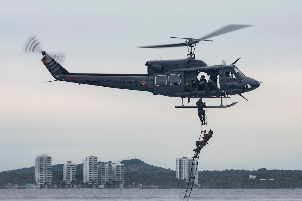 Airmen in wet suits climb out of water up a ladder into a helicopter with a city skyline in the background.