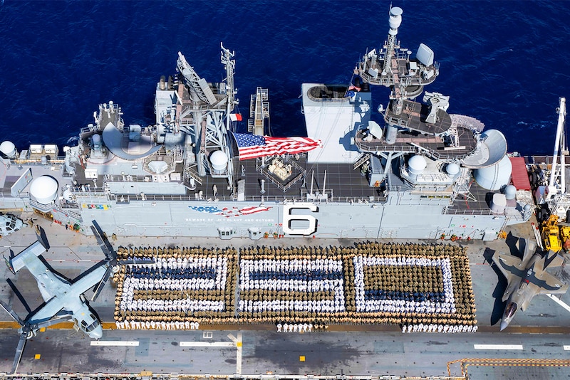 Hundreds of sailors and Marines stand in formation to form the number 250 on the flight deck of a ship at sea as seen from above.