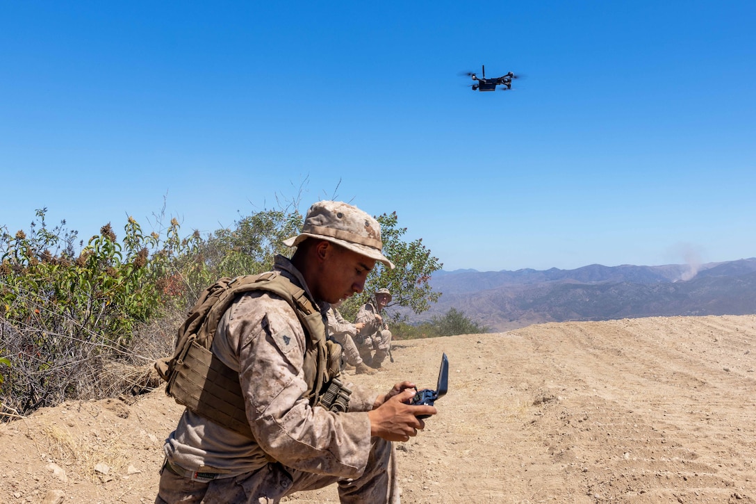A Marines kneels in the dirt while using to remote control to operate an airborne drone as fellow Marines watch during the day.